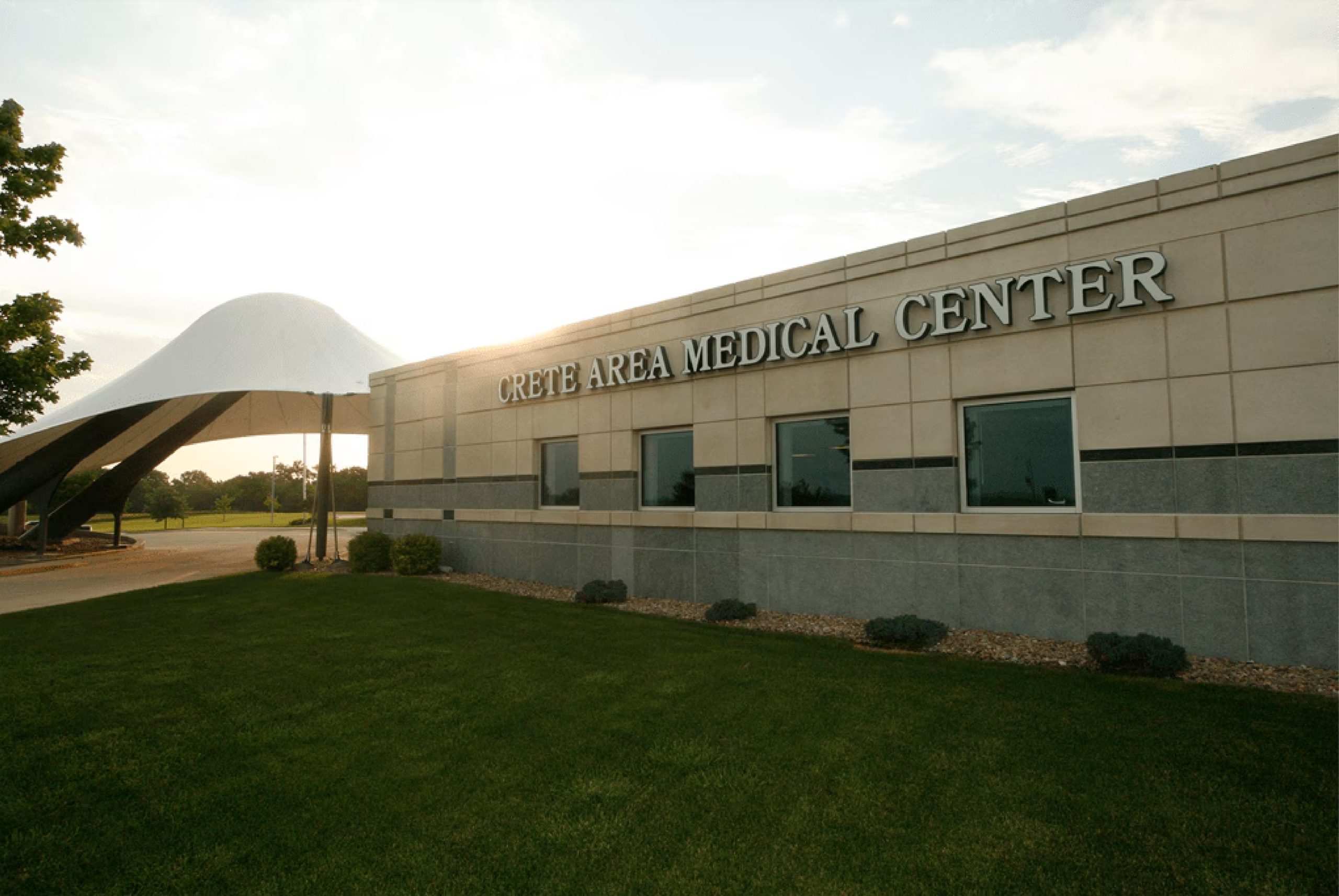 Side of Crete Area Medical Center building made of grey and tan stone, front white mushroom-shaped awning and grass in Crete, Nebraska